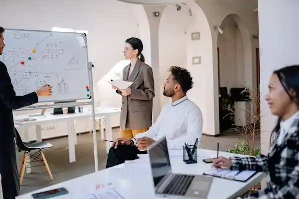 A team of professionals in a meeting room, collaborating around a whiteboard with charts and diagrams.